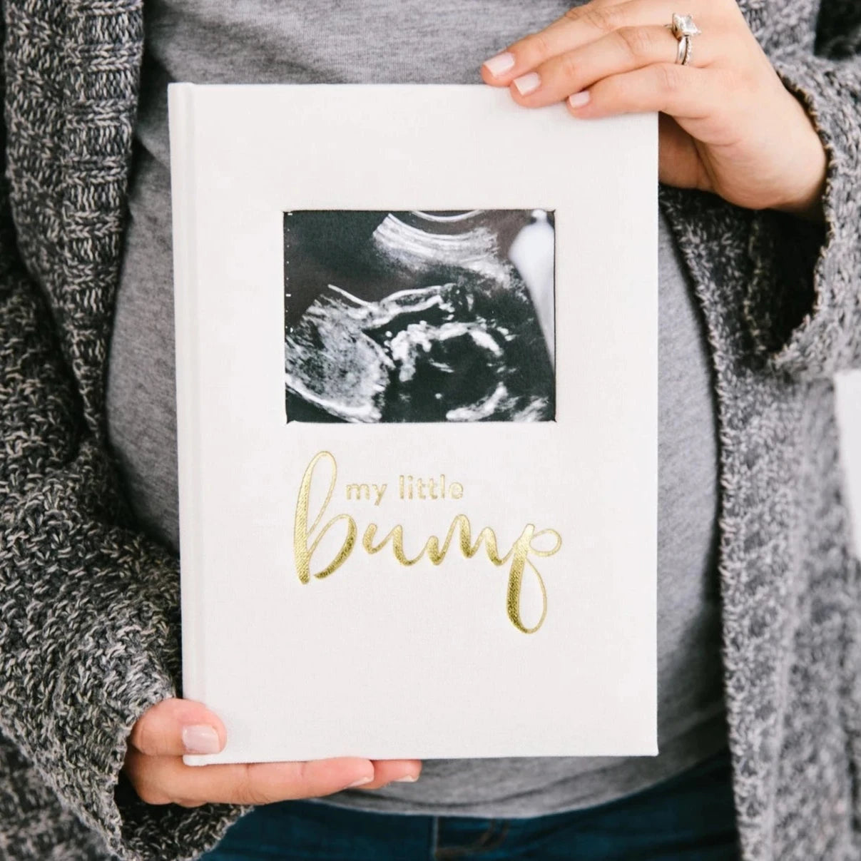 A person holding a white pregnancy journal with 'My Little Bump' printed in gold foil on the cover, featuring a photo insert of a sonogram.