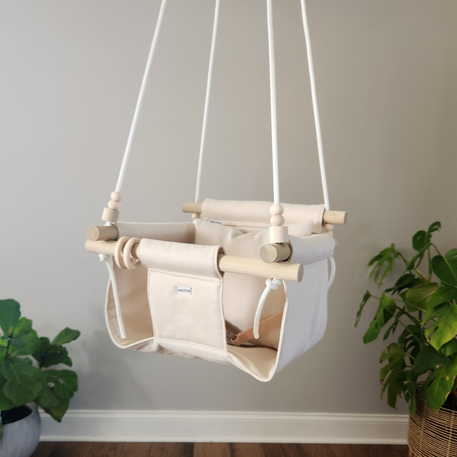 Children's swing hanging against a plain wall with plants on either side.