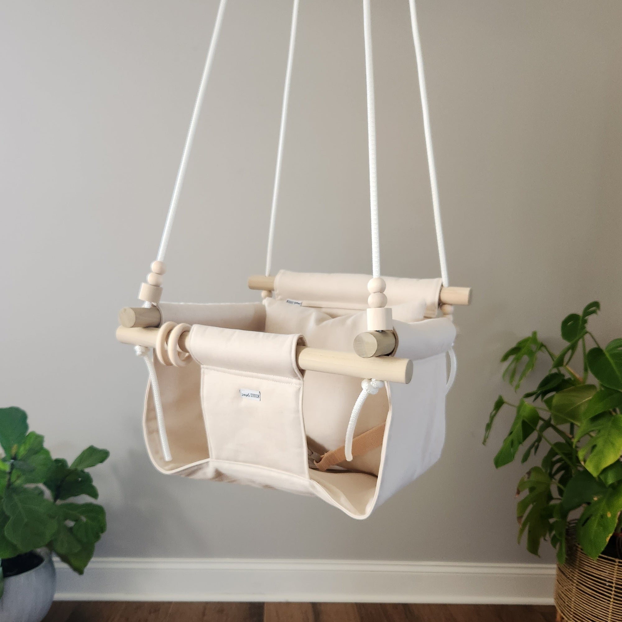 Children's swing hanging against a plain wall with plants on either side.